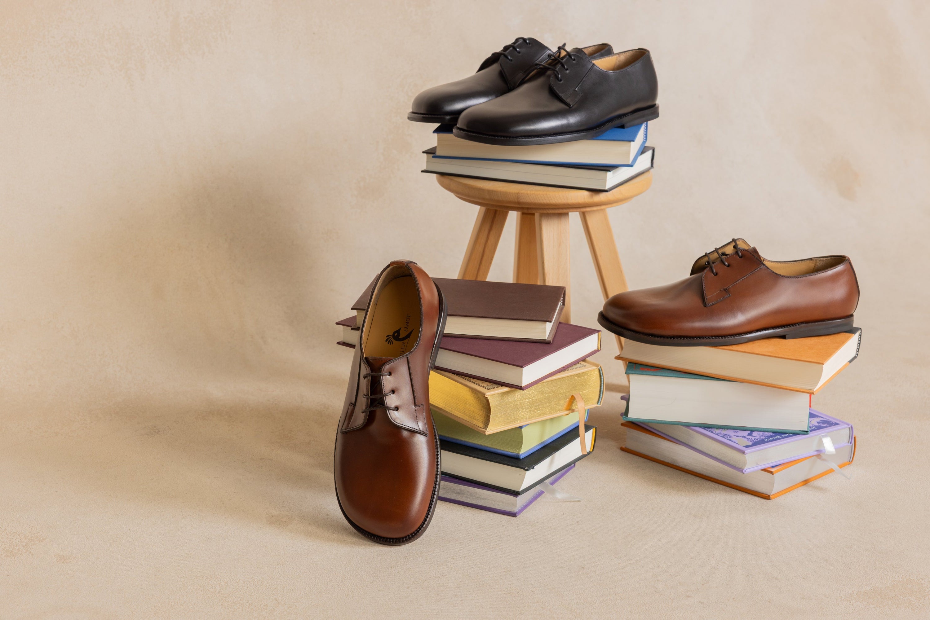 Four pairs of dress shoes arranged with stacked books on a beige studio background: two black oxford shoes on a small wooden stool, one brown oxford shoe standing upright on a book stack, and one brown lace-up shoe resting on another book stack.